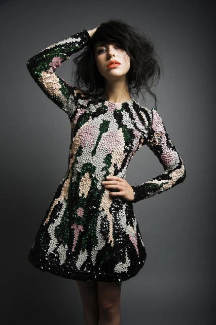 Young woman in a patterned long-sleeve dress poses against a grey backdrop, with one hand on her head and the other on her hip.