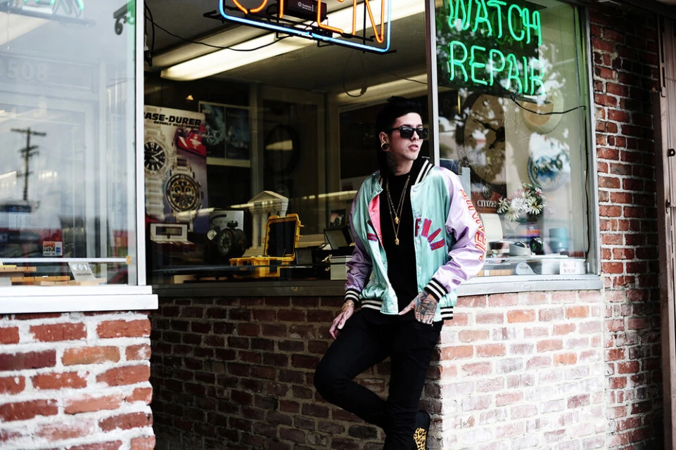 A young adult poses in front of a vintage watch repair shop, wearing a stylish jacket, sunglasses, and jewelry in an urban setting.