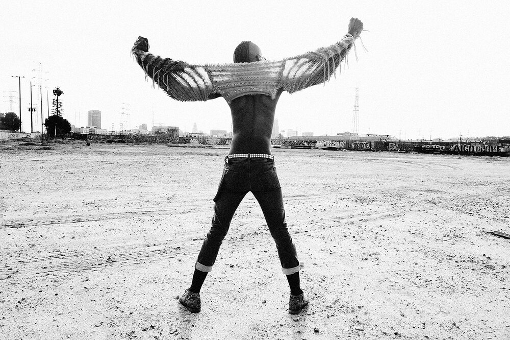 A person stands in a field, facing away from the camera, with arms raised above their head, wearing a decorative top and jeans.