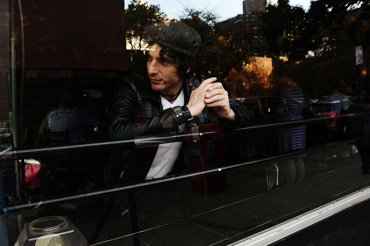 Man in leather jacket and cap looking out a window in an urban setting with blurred reflections and natural lighting indoors.