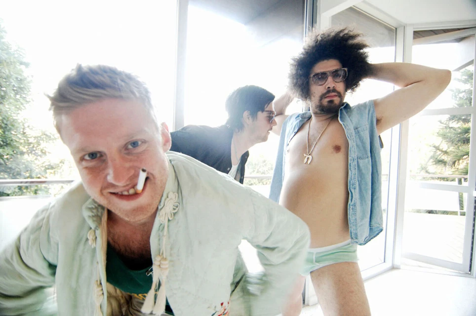 Indoor studio shot features three men: one smoking, one shirtless in underwear, and one in the background wearing sunglasses.