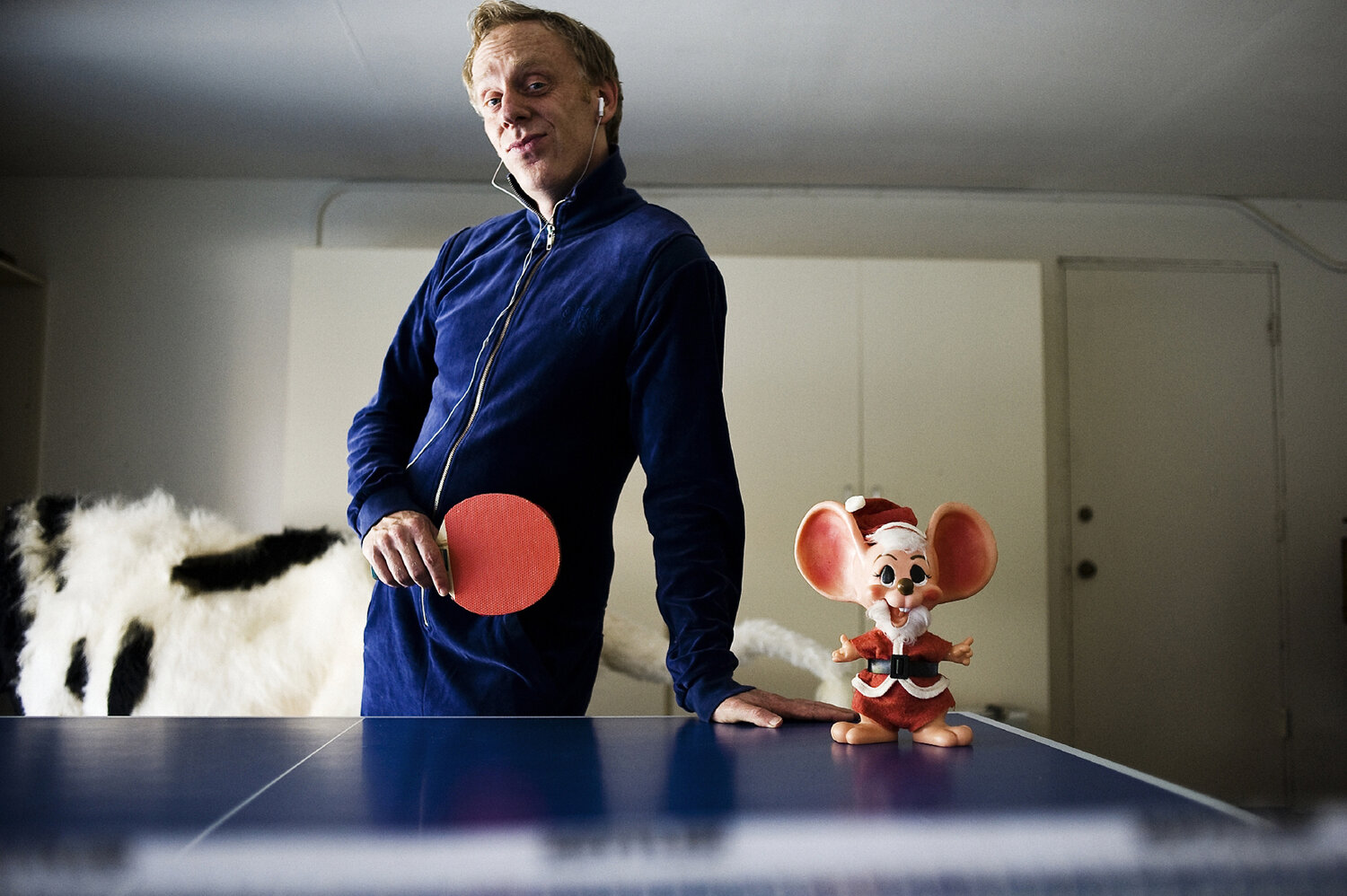 Man in blue tracksuit poses with table tennis paddle next to a vintage Mickey Mouse figurine on a ping pong table indoors.