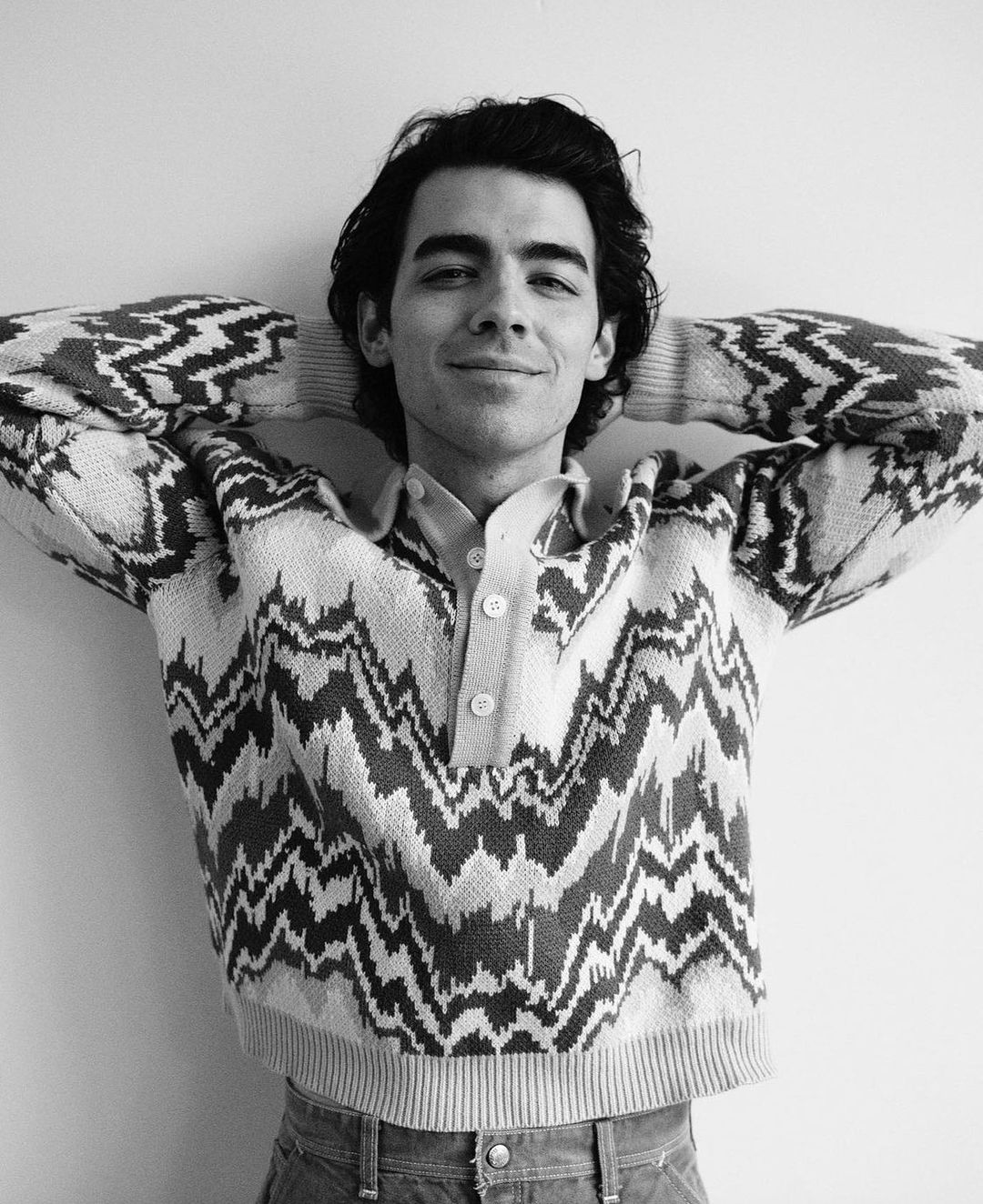 Monochrome portrait of Joe Jonas wearing a patterned knit sweater with hands behind his head against a plain white wall.