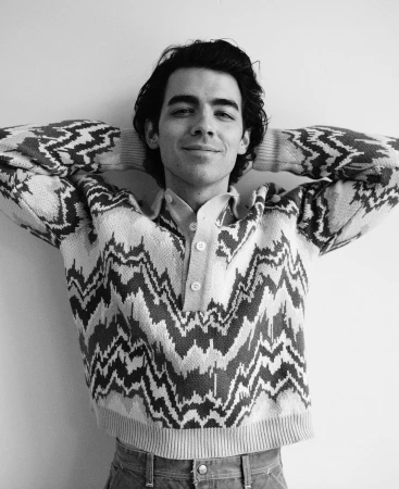 Monochrome portrait of Joe Jonas wearing a patterned knit sweater with hands behind his head against a plain white wall.