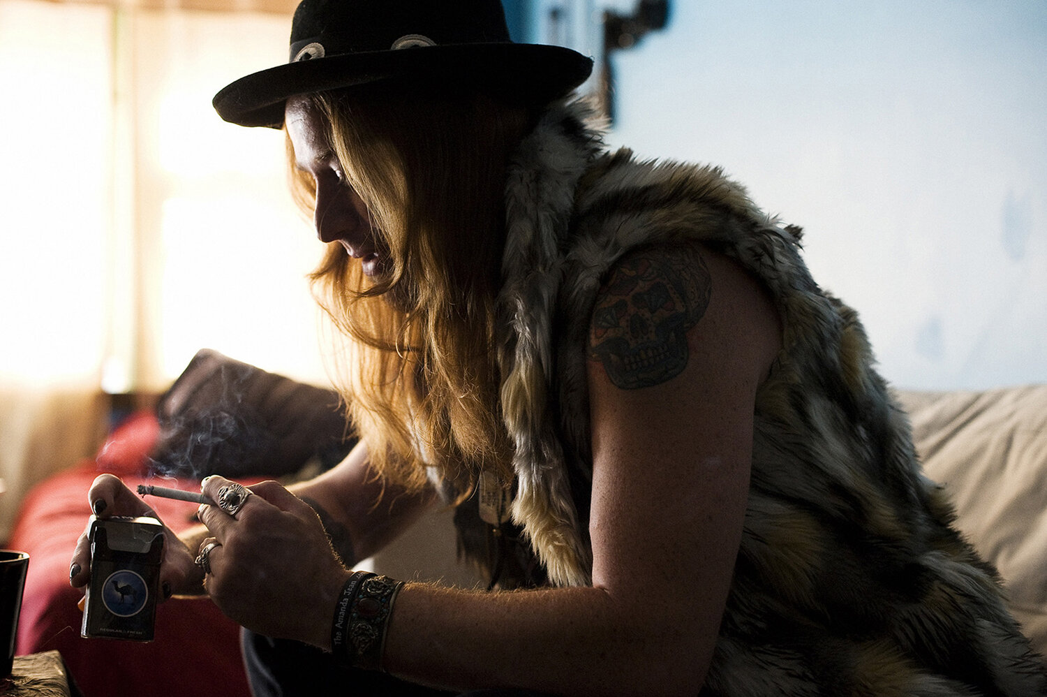 Man with long hair and hat wearing a fur vest, seated indoors, holding a lit cigarette and a lighter, looking to the left.