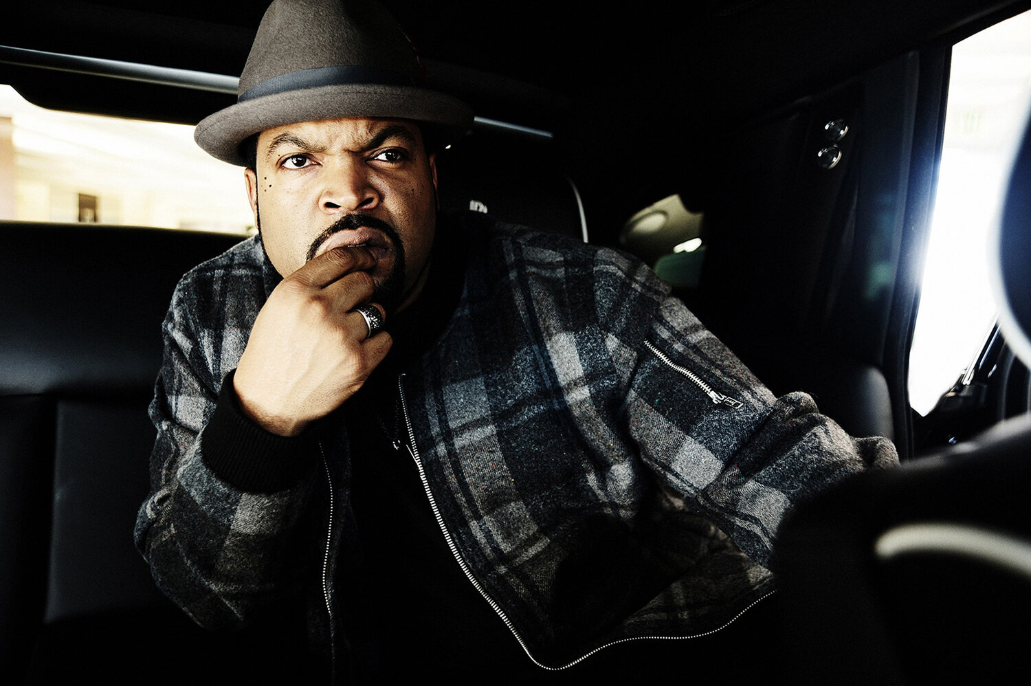 Close-up shot of Ice Cube in a car, wearing a grey plaid jacket and fedora, looking at the camera with a serious expression.