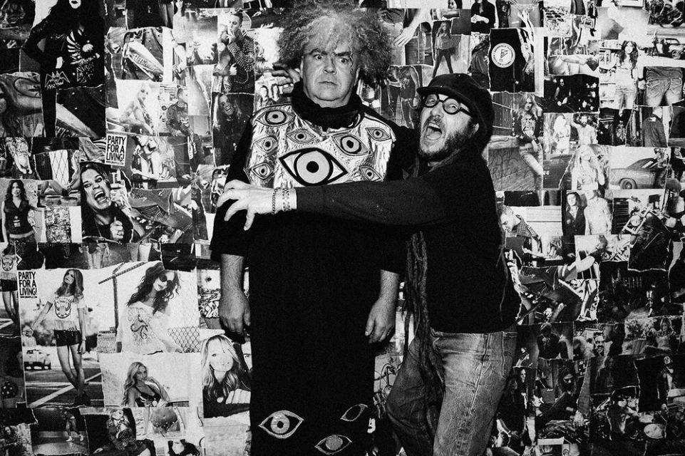 Two men pose in front of a wall covered in black and white photos; one with an afro and eye-themed shirt, and one in jeans and a hat.