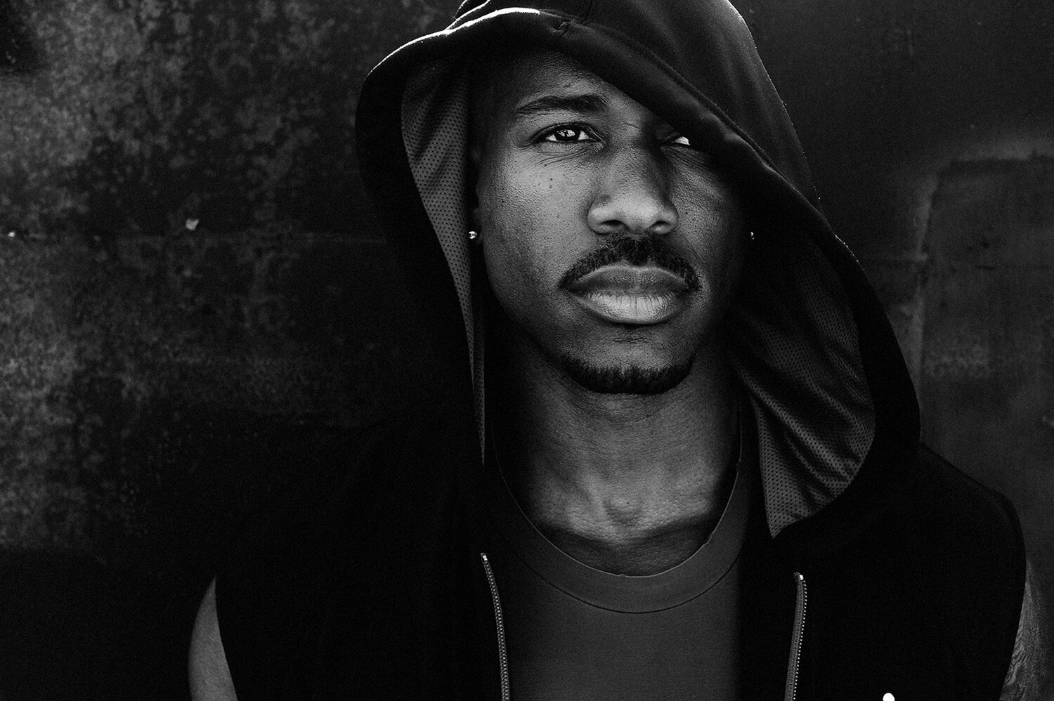Black and white portrait of an adult man in a hooded sweatshirt, his face partially shadowed, against a textured dark background.