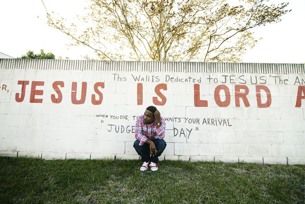 An African-American adult male crouches in front of a painted brick wall featuring red and black text, with a grass lawn foreground.