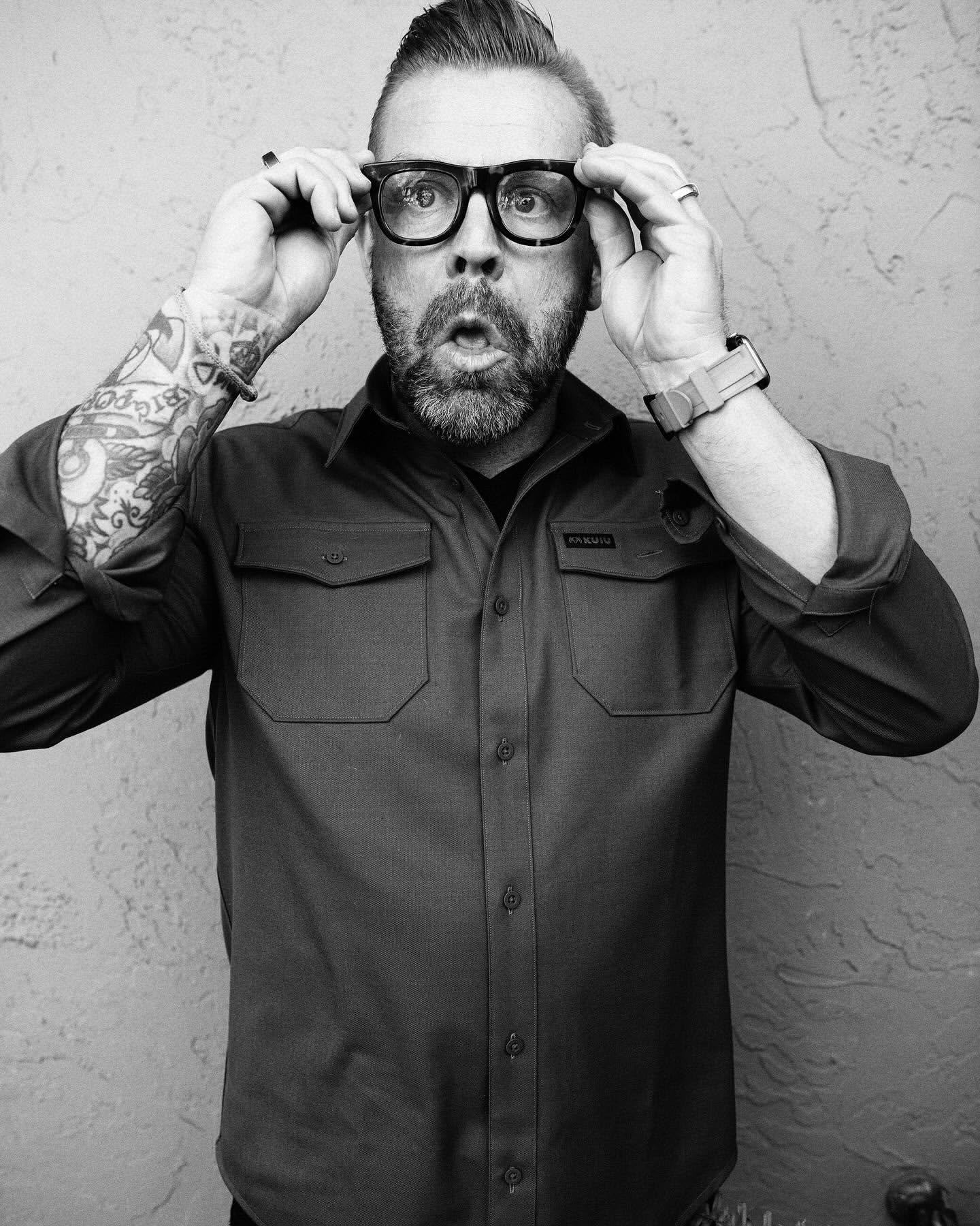 Adult male with a beard and mustache wears glasses and a button-down shirt, captured in a black and white portrait against a textured wall.
