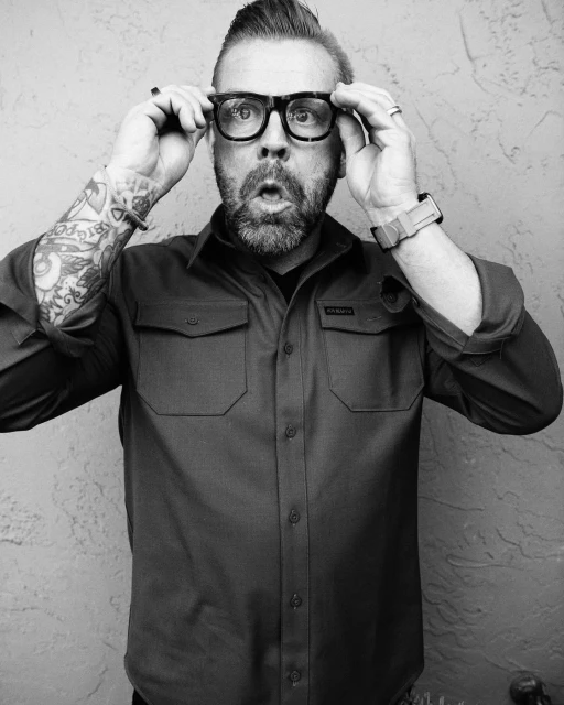 Adult male with a beard and mustache wears glasses and a button-down shirt, captured in a black and white portrait against a textured wall.