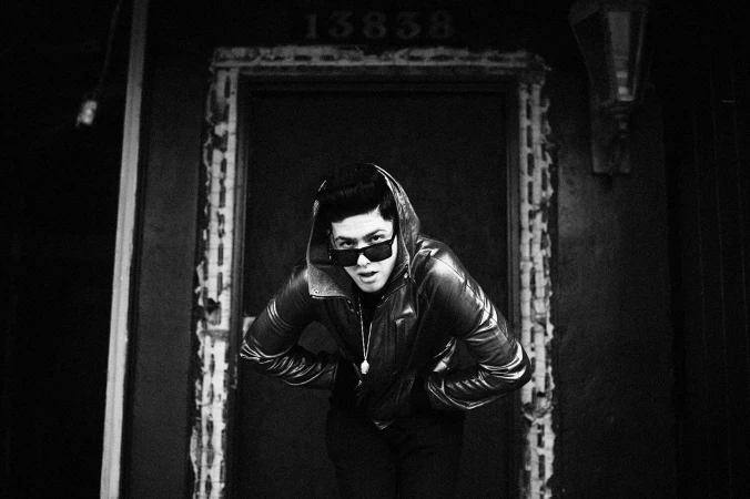 Monochrome portrait of a man wearing dark sunglasses and a hooded jacket leaning forward in a doorway by Jeff Forney.