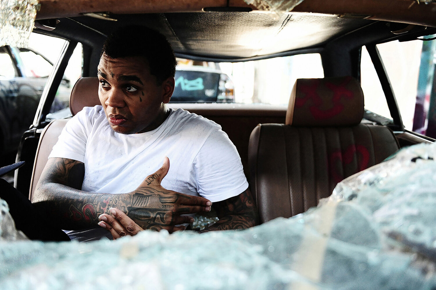 Adult male with tattoos sits in the driver's seat of a damaged car surrounded by shattered glass, creating a striking image.