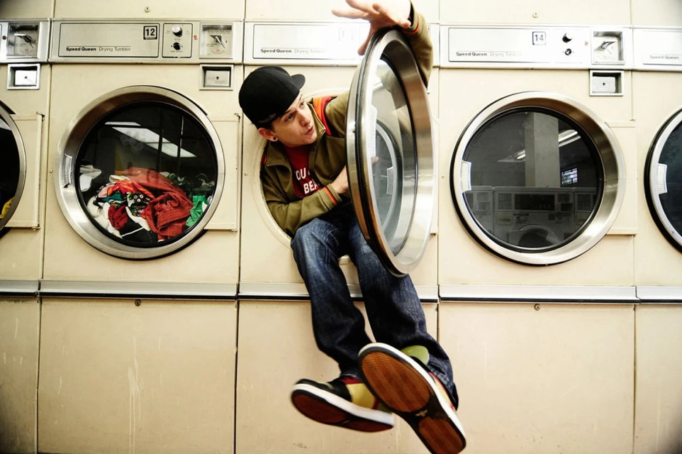 Young man in baseball cap playfully emerges from a front-loading laundromat washing machine, displaying a blend of urban style and humor.