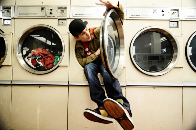 Young man in baseball cap playfully emerges from a front-loading laundromat washing machine, displaying a blend of urban style and humor.