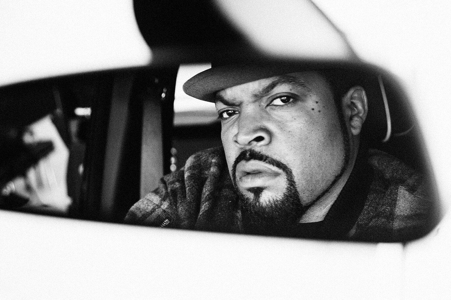 Black and white portrait of Ice Cube reflected in a car rear view mirror wearing a hat