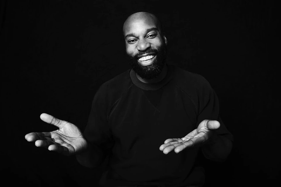 Portrait of a bearded man with dark skin, smiling and gesturing with open palms, set against a stark black background in monochrome.