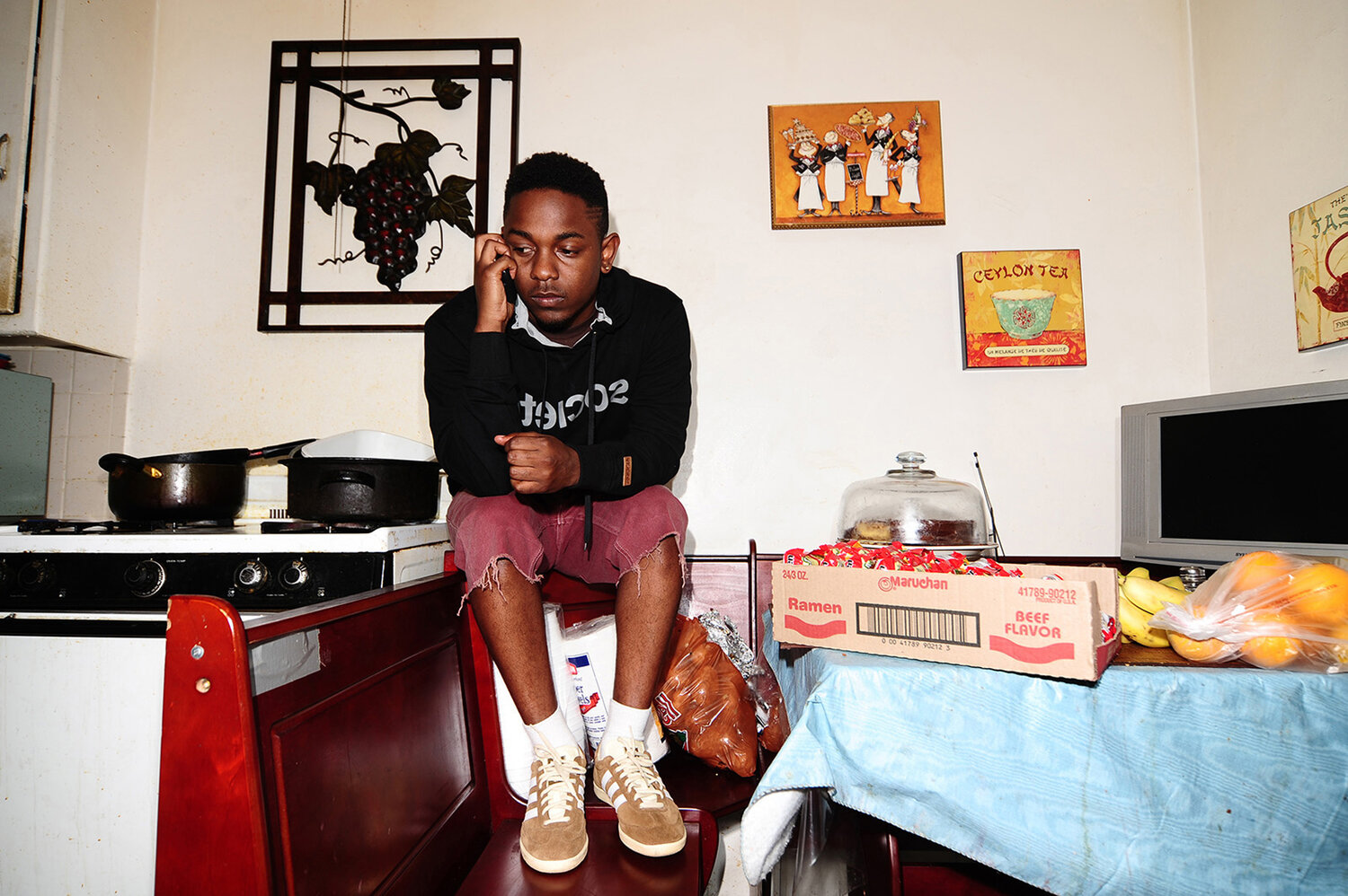 A young Black man sits on a chair in a kitchen, talking on the phone, wearing a black hoodie and red shorts, in a casual pose.