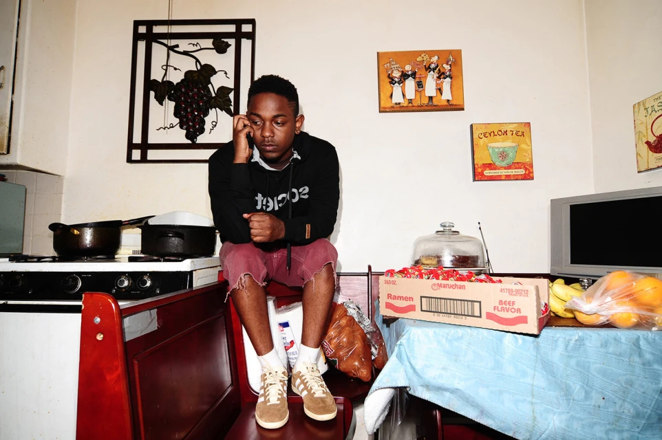 A young Black man sits on a chair in a kitchen, talking on the phone, wearing a black hoodie and red shorts, in a casual pose.