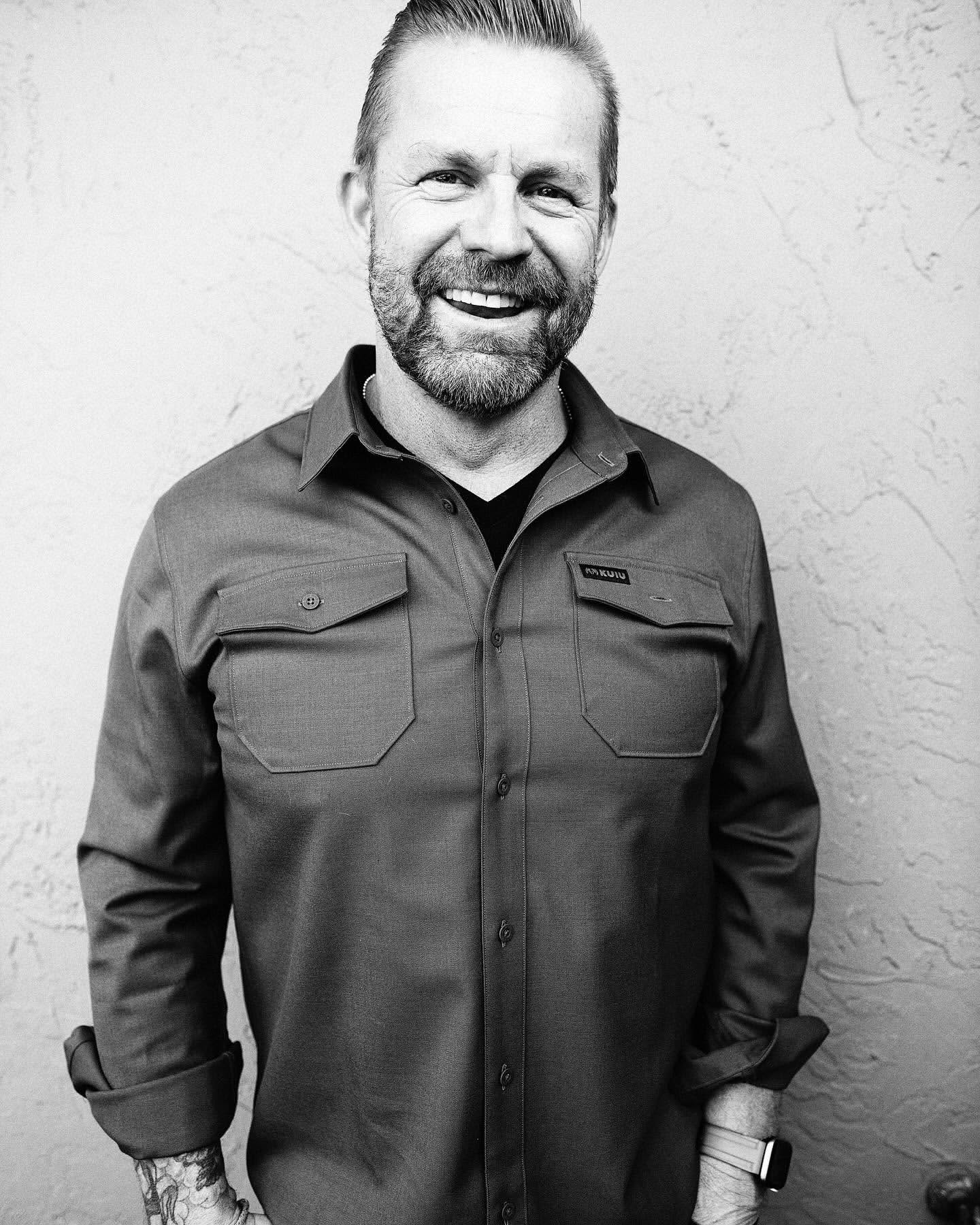 Monochrome portrait of a smiling adult man in a collared shirt with chest pockets against a textured wall backdrop.