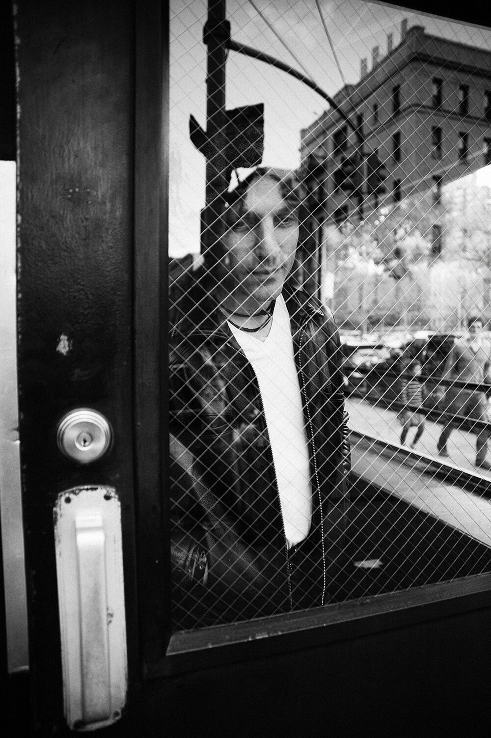 Black and white portrait of a person behind a window with a metal grid, reflecting an urban scene with buildings and pedestrians.