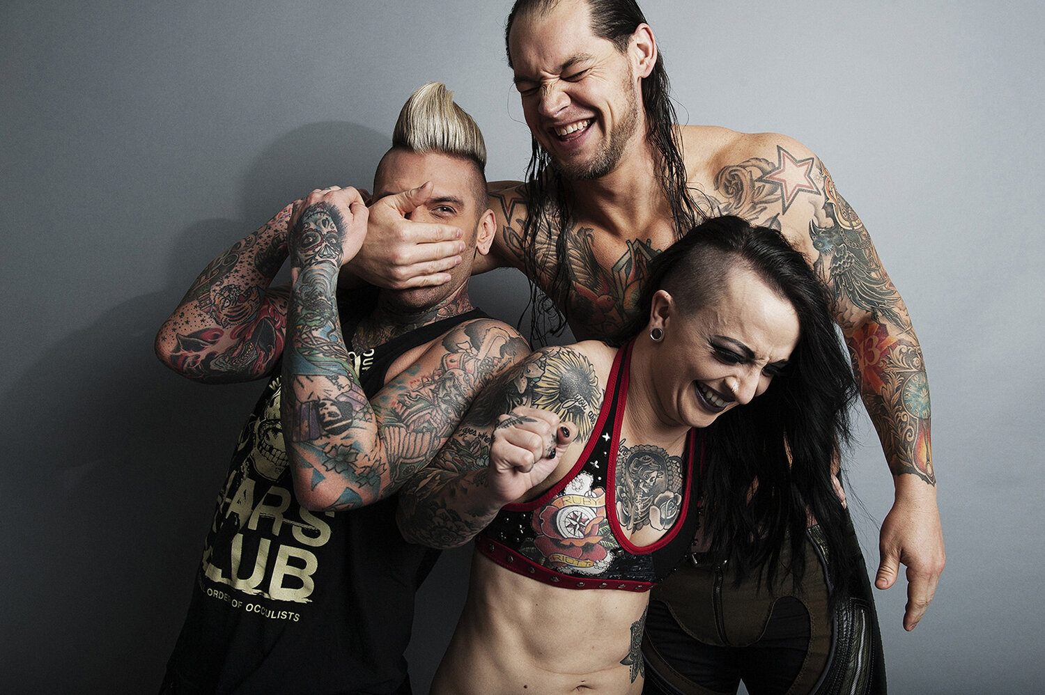 Three tattooed adults laughing and covering each other's faces against a grey background. One is shielding their eyes.