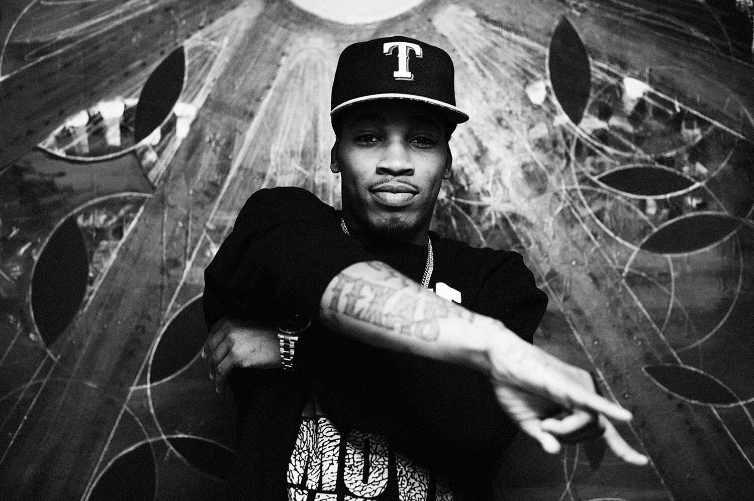Monochrome portrait of a black man wearing a baseball cap and patterned t-shirt posing in front of a textured circular art piece