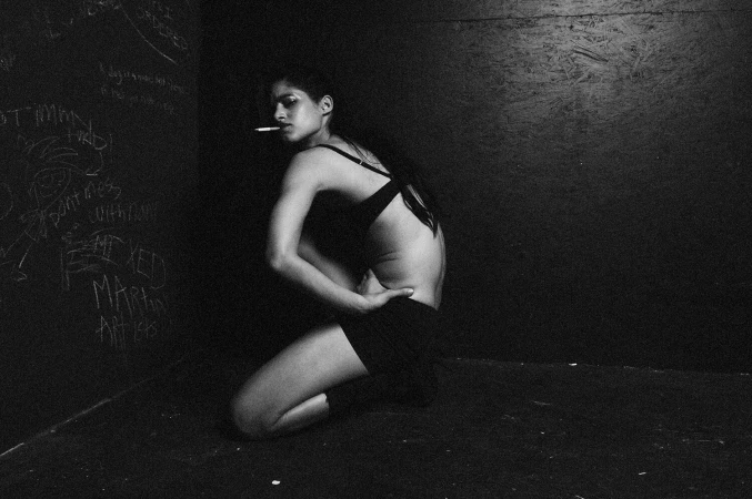 Black and white portrait of a woman crouching against a dark wall with graffiti, holding a cigarette.