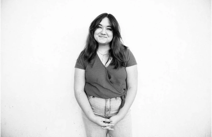 Black and white portrait of a smiling woman with long hair, wearing a wrap top and jeans, against a white backdrop.
