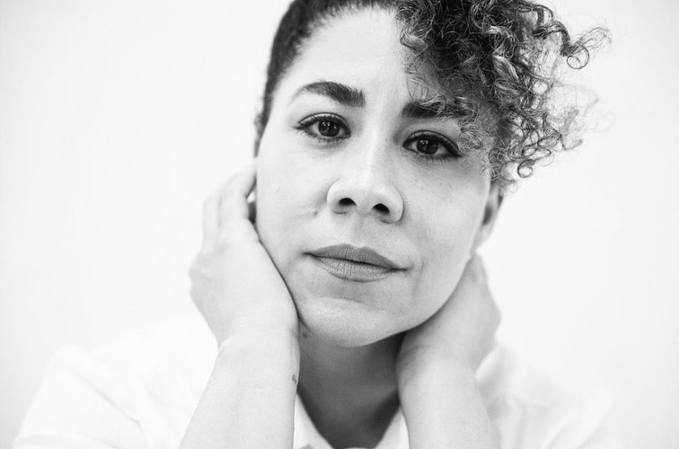 Close-up monochrome portrait of a woman with curly hair framing her face with both hands against a plain white background.