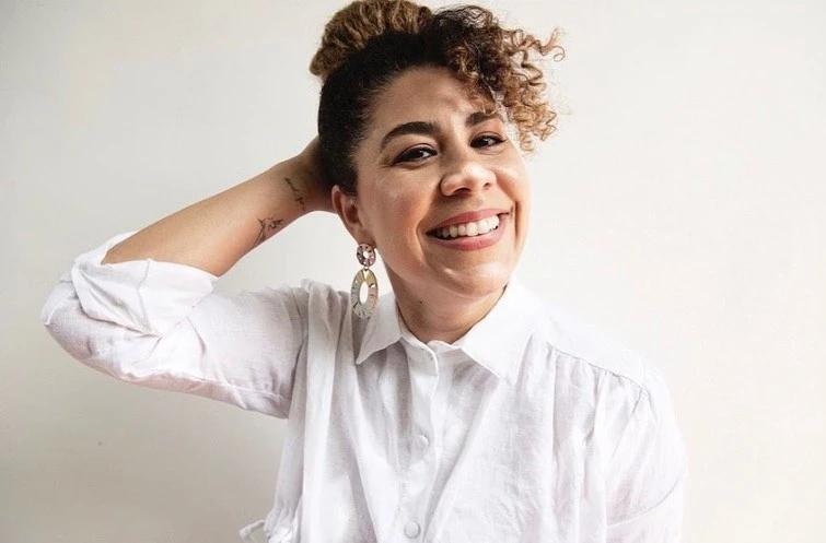 Smiling woman in a white blouse with her hand in her hair, accessorized with large earrings, posing in front of a neutral background.