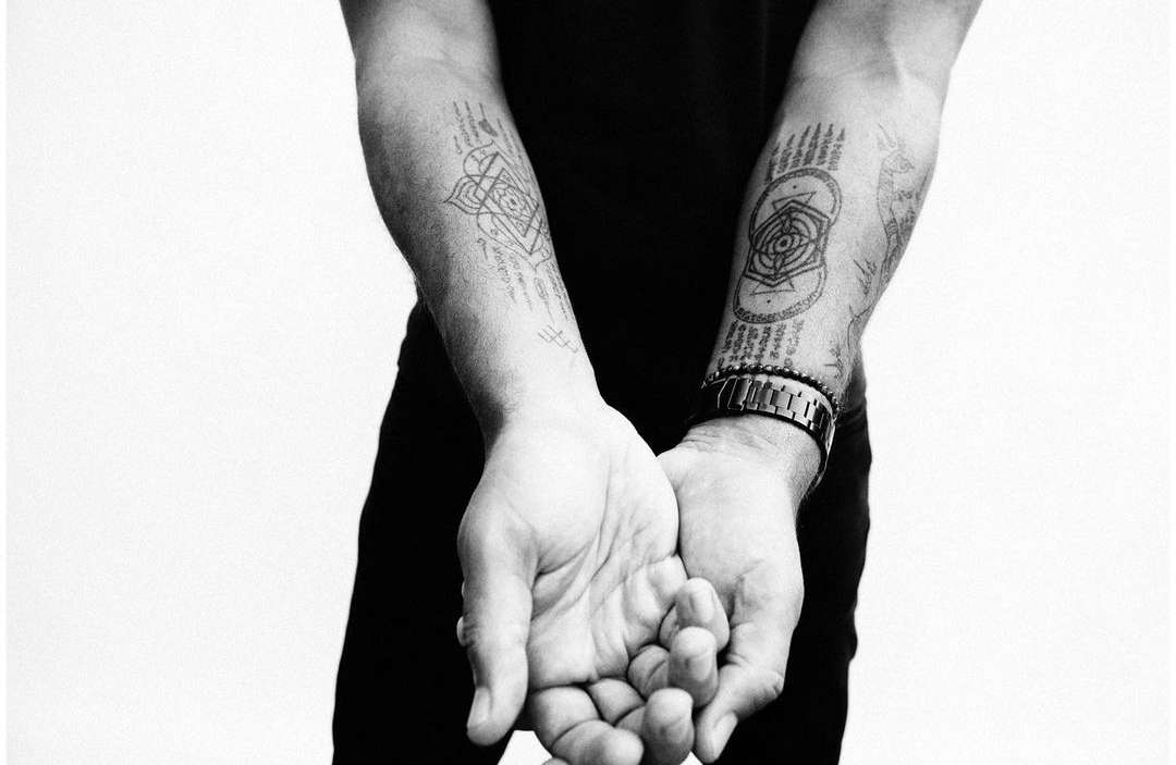 Close-up black-and-white image of a man's arms and hands, tattoos visible on his forearms, accessorized with a watch and beaded bracelets.