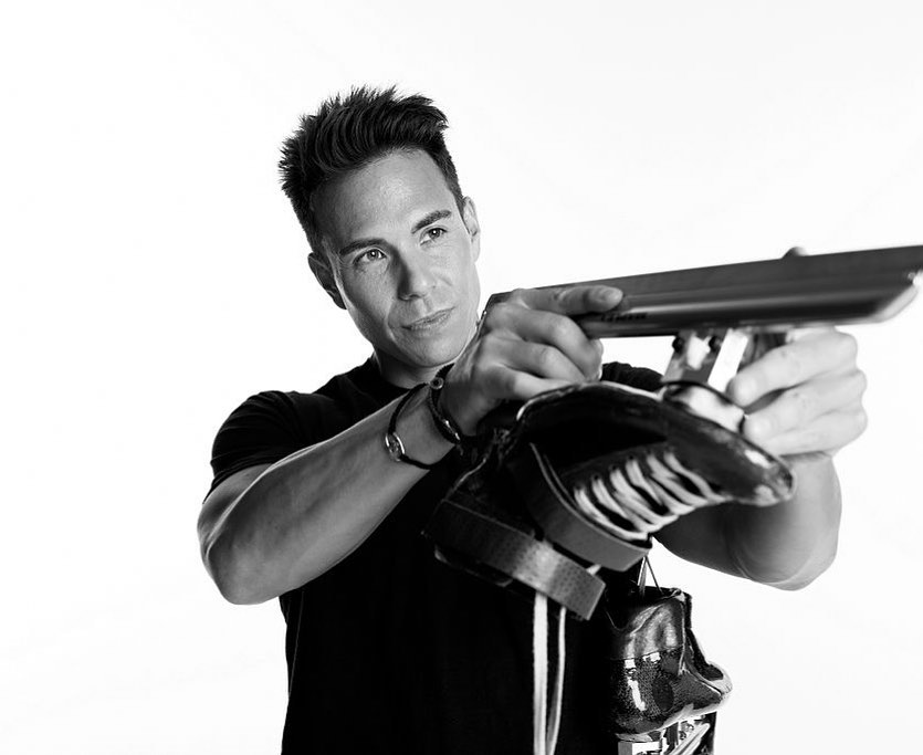 A man in a black shirt poses in a black and white studio shot holding an unknown object with both hands, looking towards the left.