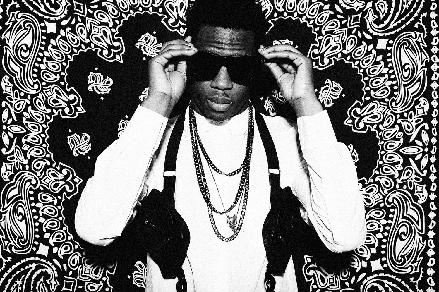 Portrait of a young adult man wearing sunglasses and chains against a patterned black and white background, showcasing street style fashion.