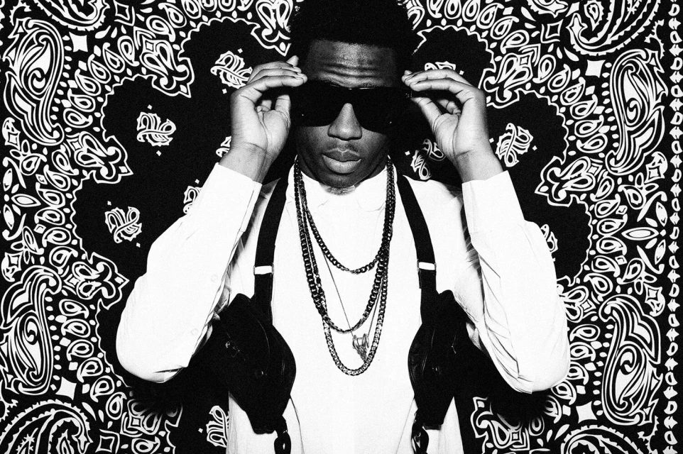 Portrait of a young adult man wearing sunglasses and chains against a patterned black and white background, showcasing street style fashion.