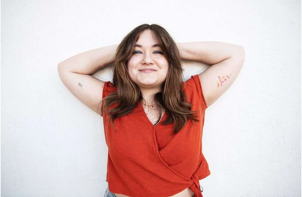 Portrait of a smiling woman with brown hair and tattoos on her arms, wearing a red wrap top in front of a white wall.