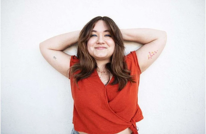 Portrait of a smiling woman in a rust orange wrap top with arms raised against a plain white wall, photographed by Jeff Forney.