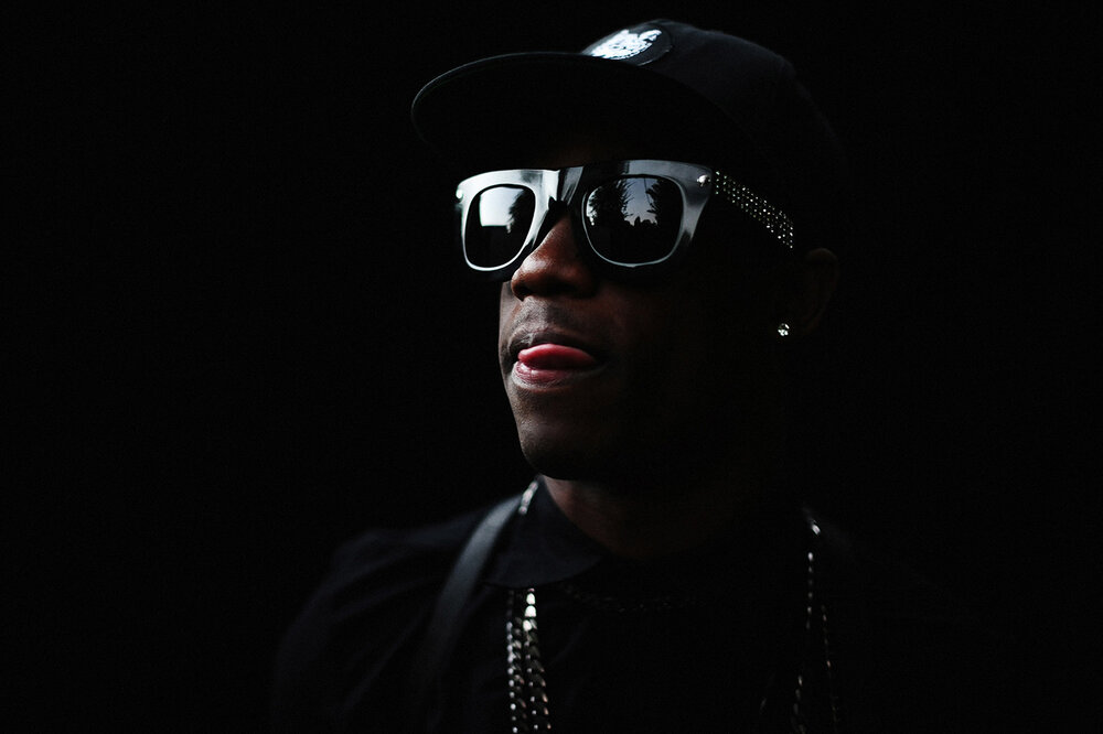 Close-up portrait of an African-American adult male wearing a black baseball cap and reflective sunglasses against a dark background.