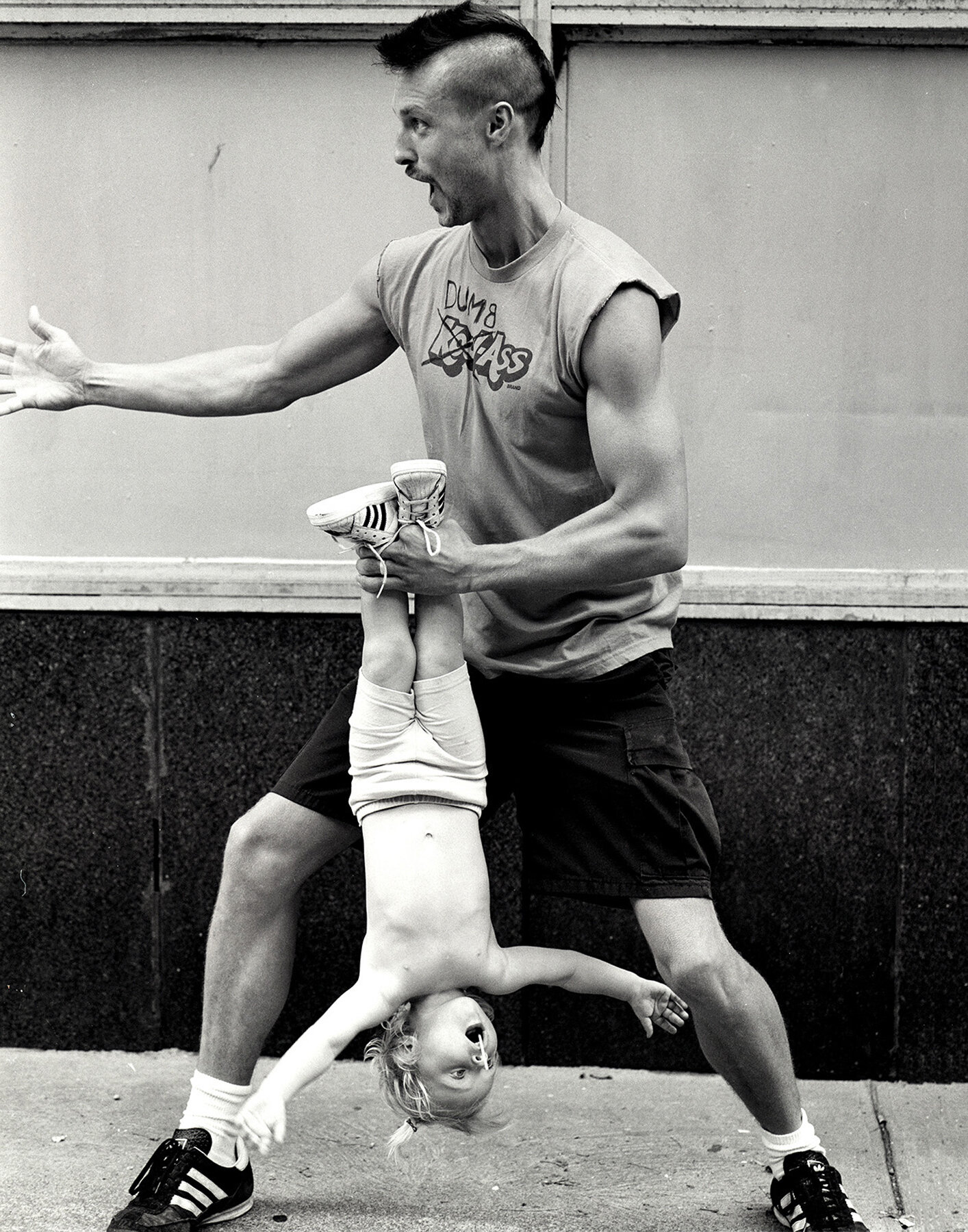 Black and white shot of a man holding a young child upside down by the feet; the man has a mohawk and open mouth, outside.