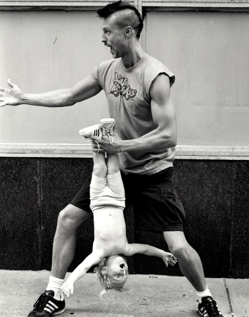 Black and white shot of a man holding a young child upside down by the feet; the man has a mohawk and open mouth, outside.