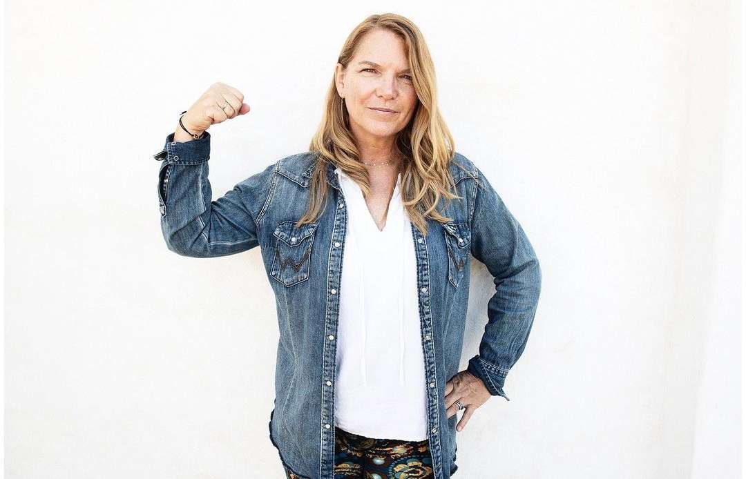 Portrait of an adult woman in a denim jacket flexing her arm against a white background, showcasing a casual and confident pose.