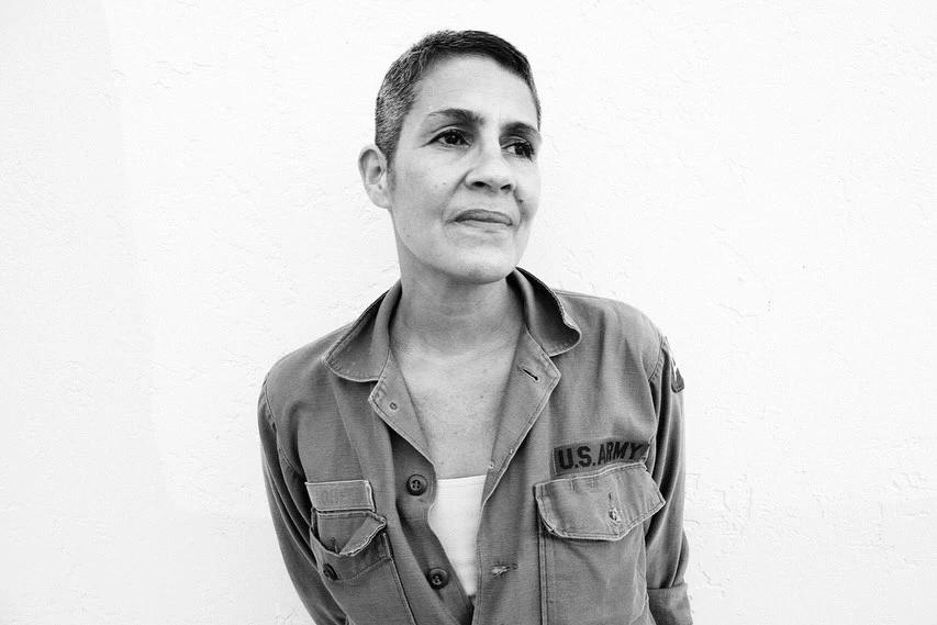 Monochrome portrait of an adult woman with short hair, wearing an unbuttoned US Army jacket in front of a textured white wall.