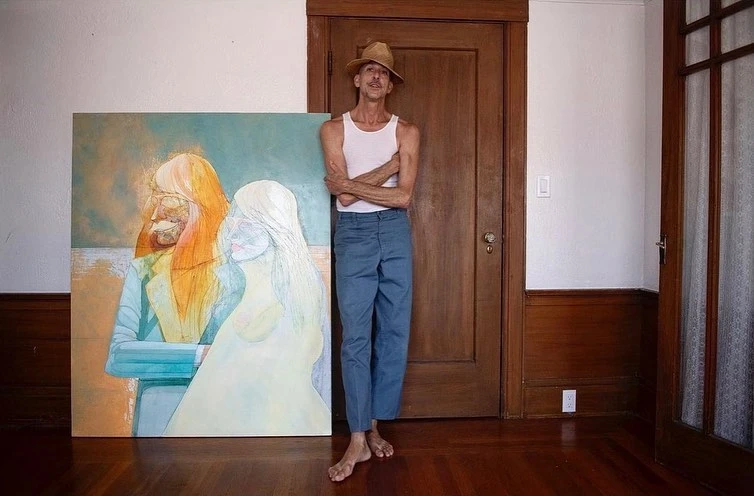 Man in tank top and trousers standing next to a canvas painting in a room with hardwood floors, dark wood trim, and a tan wall.