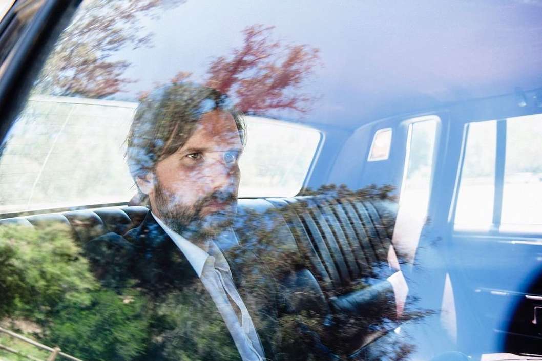 Man in a suit rides in a car, reflected in the window, trees pass by, creating an outdoor, nature-infused, and travel-focused image.