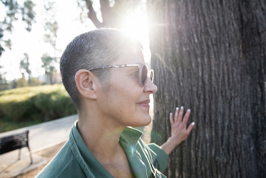 Adult woman in sunglasses leans against tree trunk in sun-drenched park setting, wearing a green shirt with a happy expression.