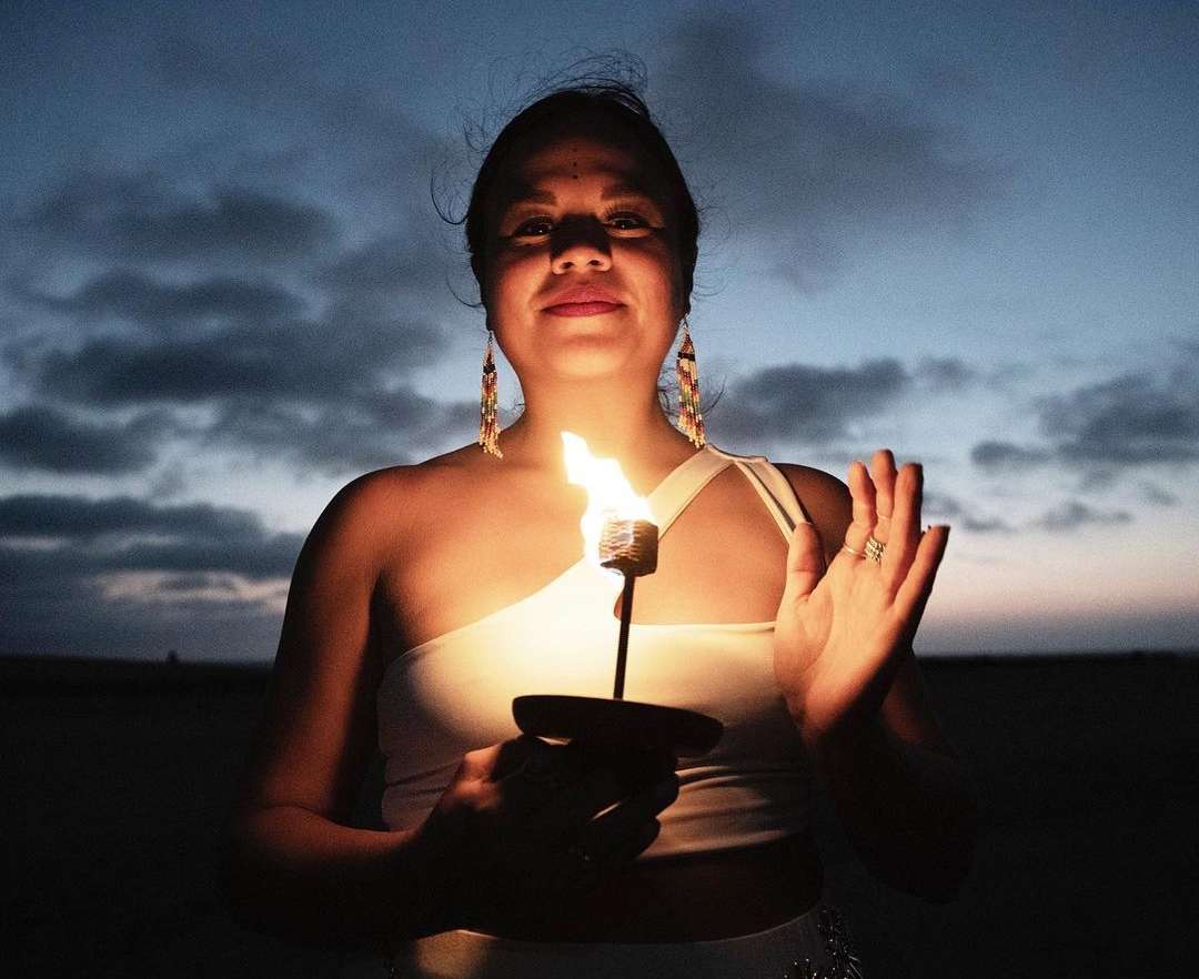 Woman holding a burning torch against a twilight sky. She is wearing a white, one-shoulder top and ornate earrings. The sky is dark.