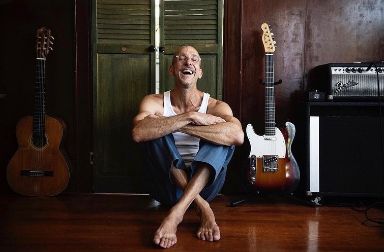 An adult male sitting barefoot on a wooden floor with two guitars and a Fender amplifier inside a room with wooden doors.