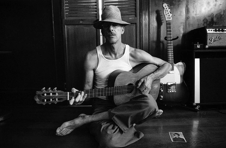 Black and white photo of a seated man playing guitar with a Fender guitar, amp, and small photo in the background.