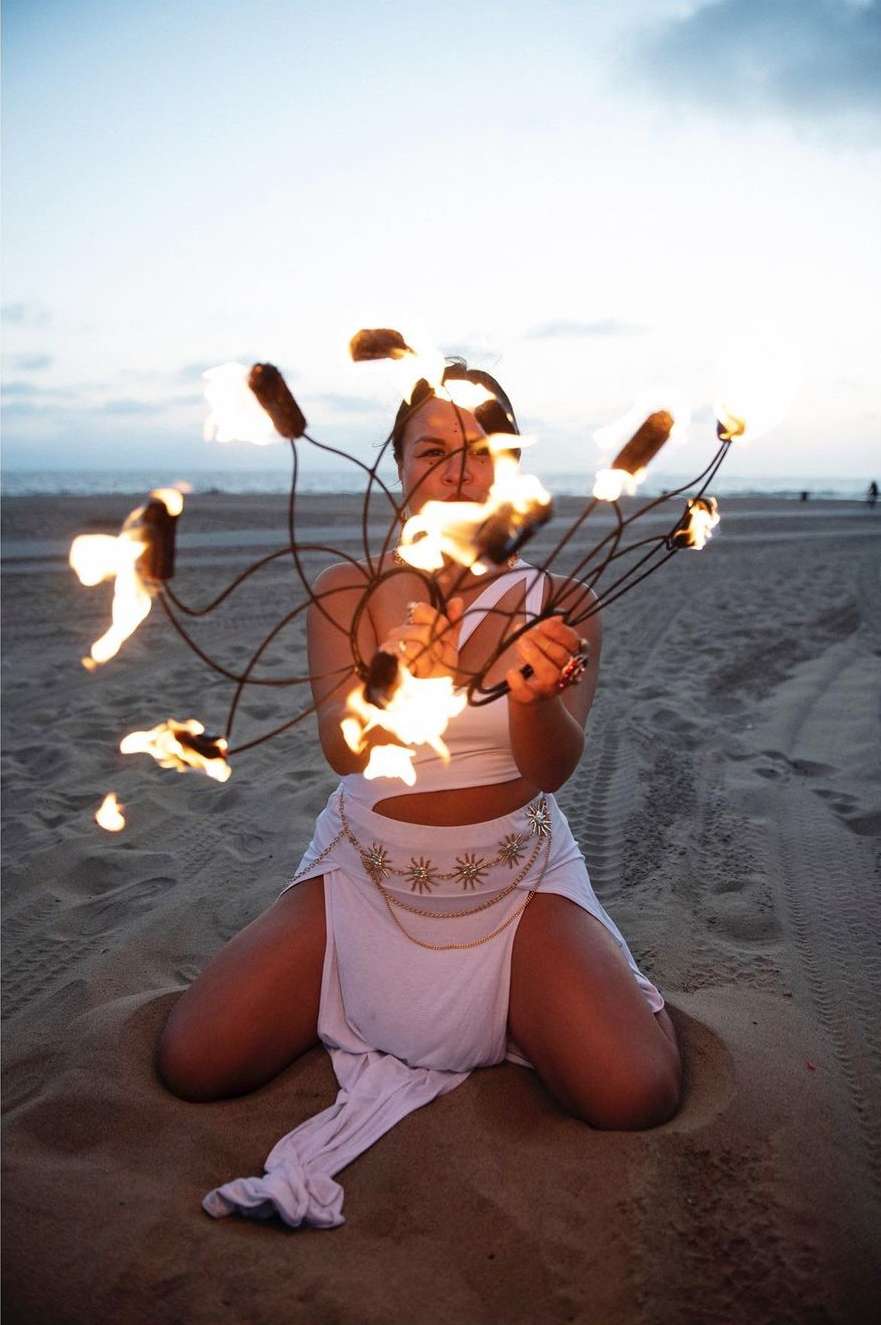 Woman in white outfit on sand holds burning fire sticks during a performance at sunset on a beach with ocean and sky background.