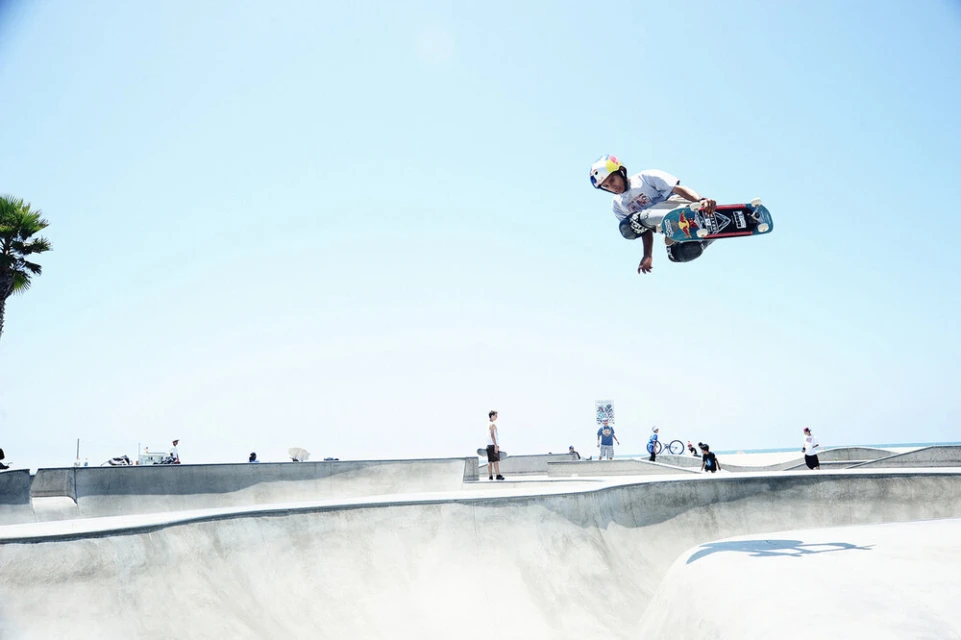 A skateboarder performs a trick mid-air at a skatepark on a sunny day. The skateboarder is wearing a helmet and safety pads.